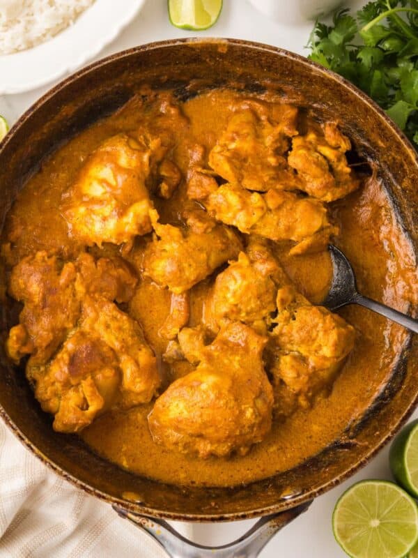 A skillet filled with chicken curry with chicken thighs coated in thick, orange sauce. Sliced limes, fresh cilantro, a bowl of white rice, and a beige towel surround the skillet on a white surface.