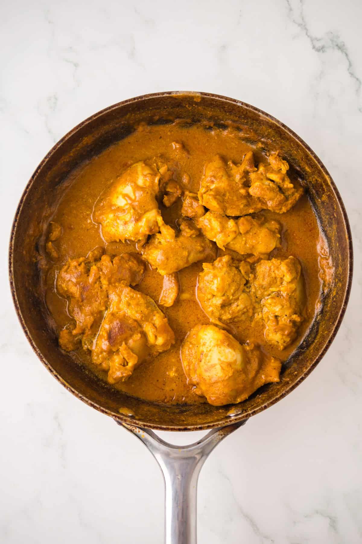 A frying pan filled with pieces of chicken cooked in a rich, orange-colored curry sauce, placed on a white marble surface.
