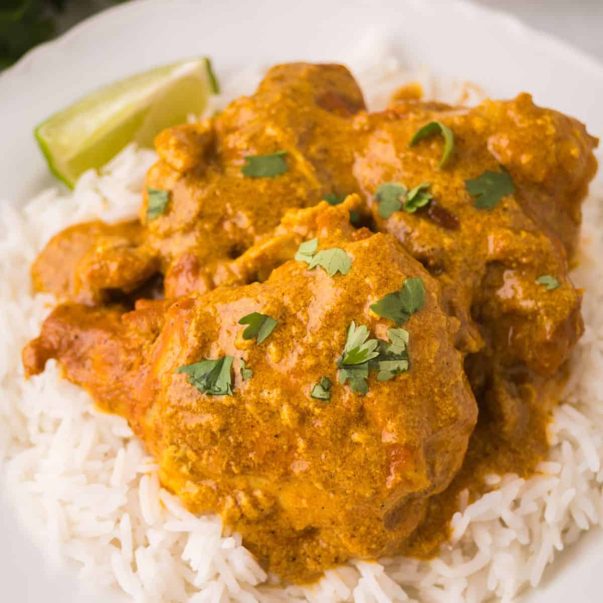 A plate of white rice topped with creamy orange chicken curry made with tender chicken thighs, garnished with chopped cilantro and served with a lime wedge.