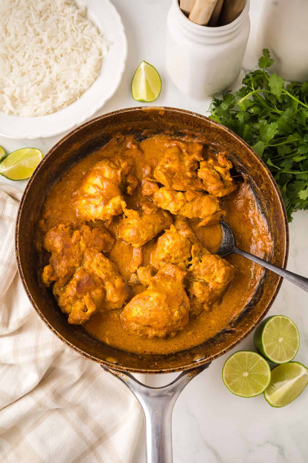 A skillet filled with creamy orange chicken curry, surrounded by lime slices, fresh cilantro, a white bowl of rice, and a white towel on a marble surface. A spoon rests inside the skillet.