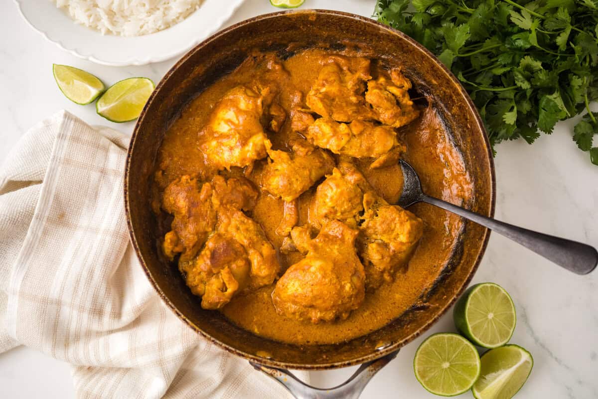 A bowl of chicken tikka masala in creamy orange sauce with a spoon, surrounded by fresh cilantro, lime wedges, and a plate of white rice on a white surface.