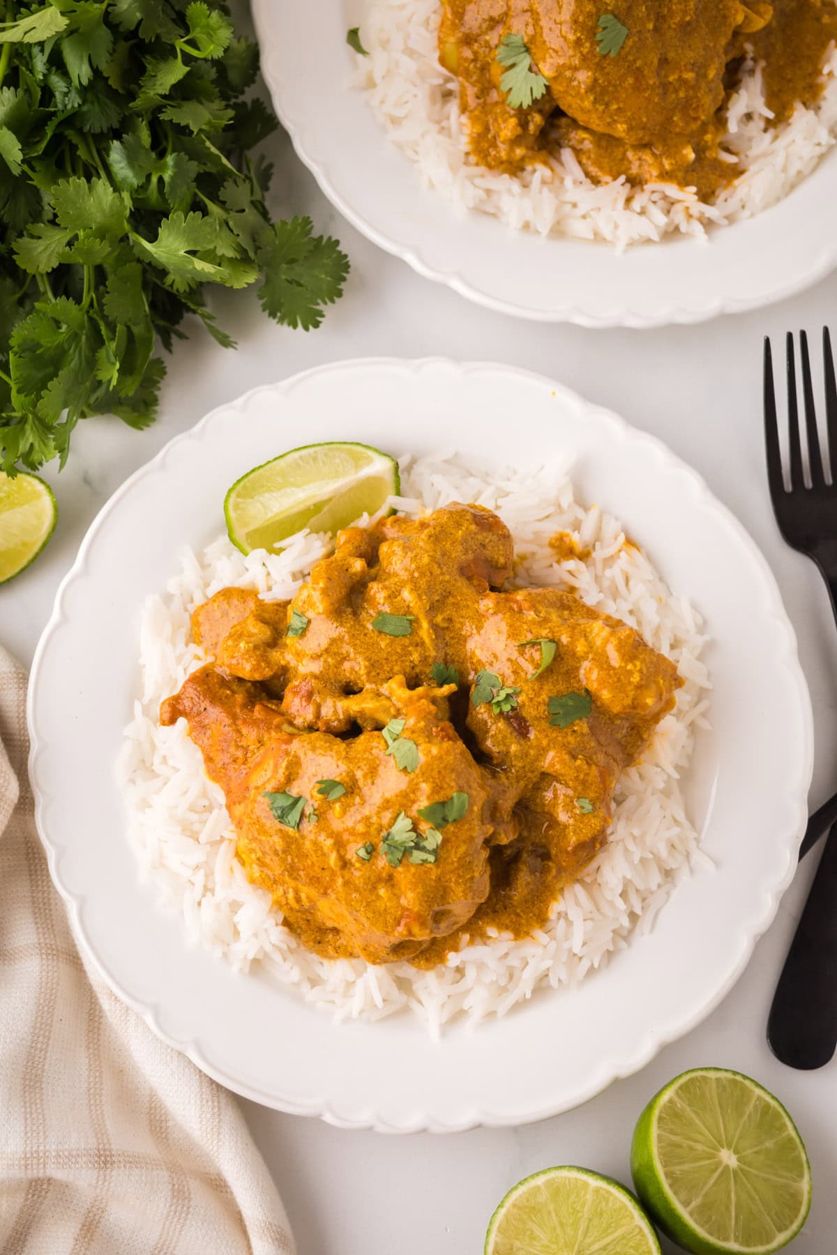 A white plate with chicken curry garnished with cilantro, served over white rice, with lime wedges on the side. Fresh cilantro, a fork, and another plate of curry are nearby.