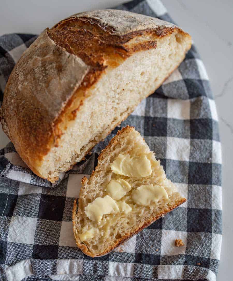 A loaf of rustic bread rests on a black and white checkered cloth, with a slice in front spread with melted butter.