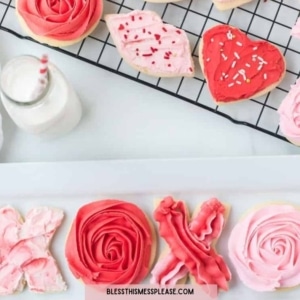 A variety of Valentine’s Day sugar cookies decorated with pink, red, and white icing—including hearts, roses, and XOXO—are displayed on a cooling rack and plate next to milk, making a sweet treat alongside your Valentine’s Day dinner suggestions.