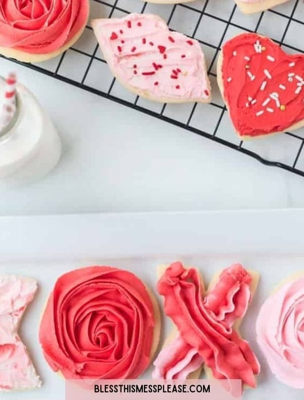 A variety of Valentine&rsquo;s Day sugar cookies decorated with pink, red, and white icing&mdash;including hearts, roses, and XOXO&mdash;are displayed on a cooling rack and plate next to milk, making a sweet treat alongside your Valentine&rsquo;s Day dinner suggestions.