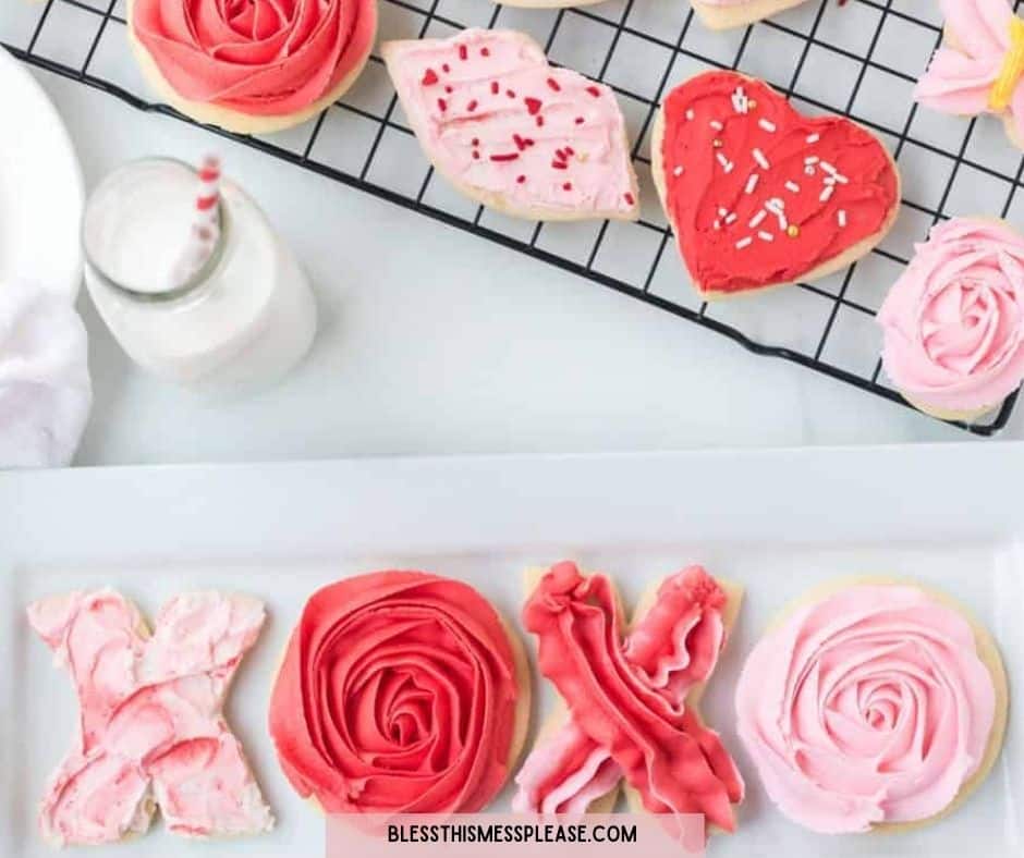 A variety of Valentine’s Day sugar cookies decorated with pink, red, and white icing—including hearts, roses, and XOXO—are displayed on a cooling rack and plate next to milk, making a sweet treat alongside your Valentine’s Day dinner suggestions.