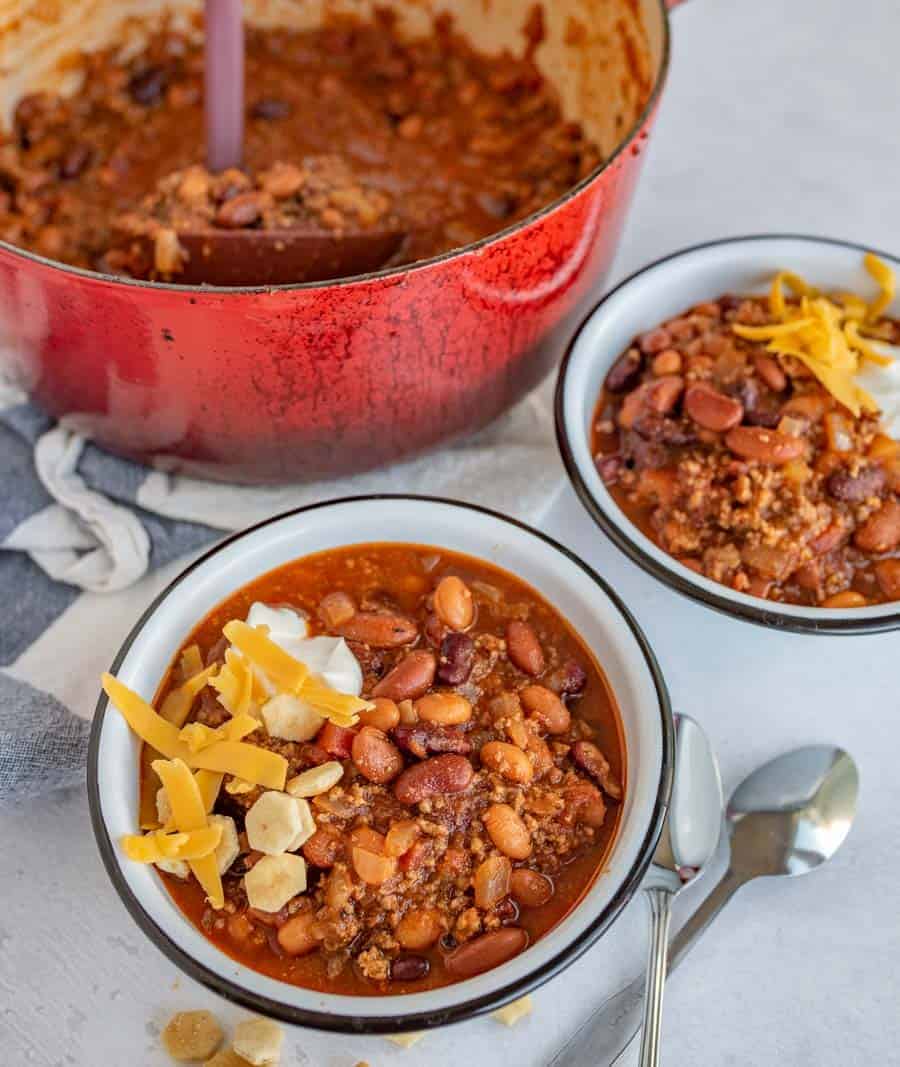 Two bowls of chili topped with shredded cheese and oyster crackers sit beside a red pot filled with more chili. Wondering what to serve with chili? A spoon and napkin are placed next to the bowls on a white surface.