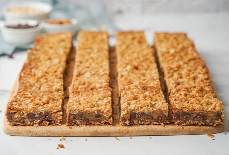 Six rectangular oat bars with visible chocolate chips are arranged neatly on a wooden cutting board. The background is blurred, showing bowls with ingredients on a white surface.