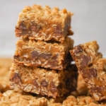 Three stacked oatmeal bars filled with gooey caramel and chocolate chips, with an additional bar leaning against the stack. The texture appears crumbly and rich, with golden brown edges and visible oats.