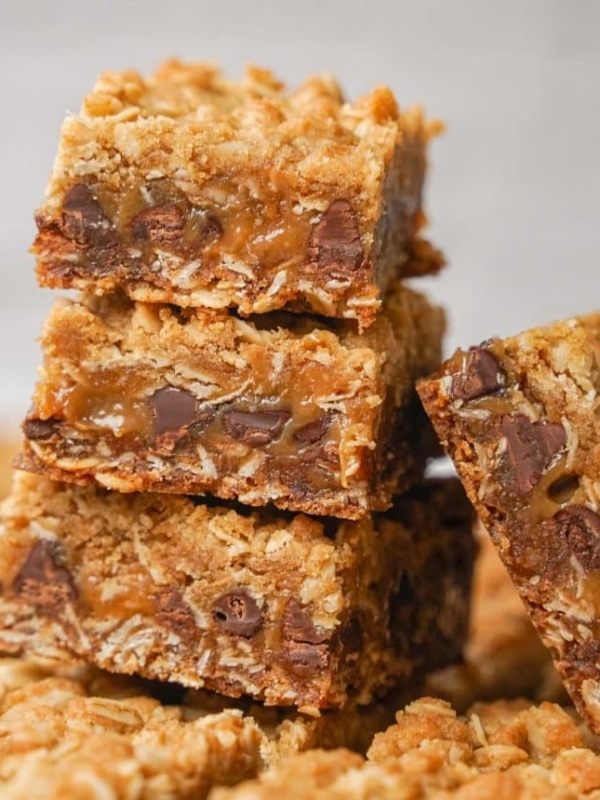 Three stacked oatmeal bars filled with gooey caramel and chocolate chips, with an additional bar leaning against the stack. The texture appears crumbly and rich, with golden brown edges and visible oats.