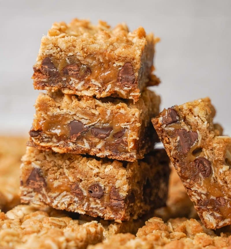 Three stacked oatmeal bars filled with gooey caramel and chocolate chips, with an additional bar leaning against the stack. The texture appears crumbly and rich, with golden brown edges and visible oats.