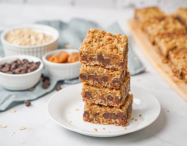 Three oatmeal chocolate chip bars stacked on a white plate, with bowls of oats, chocolate chips, and peanut butter in the background on a light surface. More bars are visible on a wooden board.