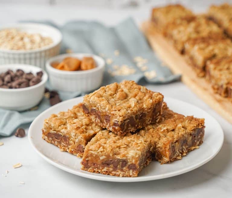 Three oatmeal caramel chocolate bars are stacked on a white plate, with more bars, chocolate chips, oats, and caramel bits in small bowls blurred in the background.