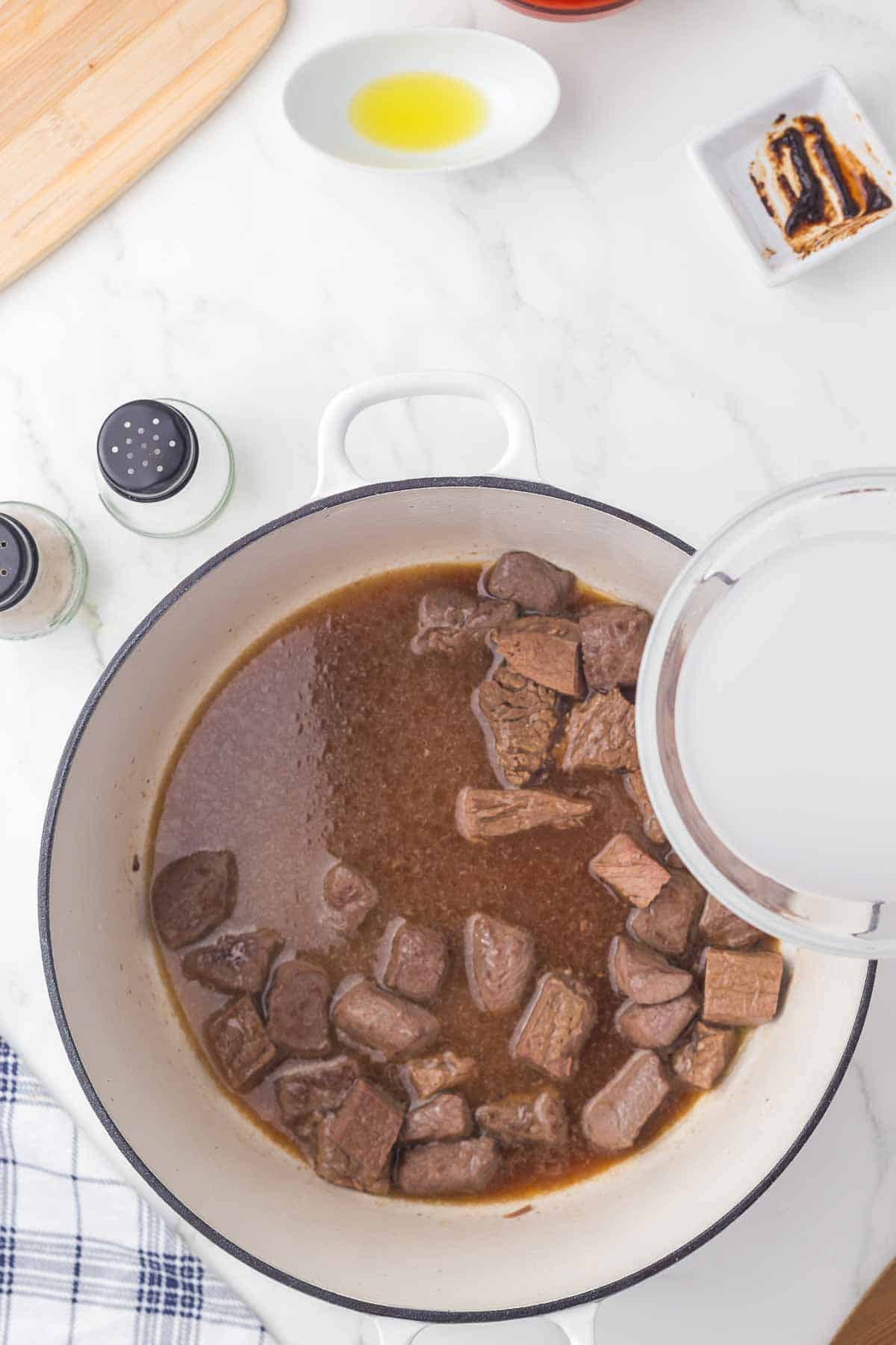 Chunks of beef simmer in broth inside a white pot on a marble countertop, surrounded by seasonings, a small dish of oil, a slice of bread, a cutting board, and a striped kitchen towel.