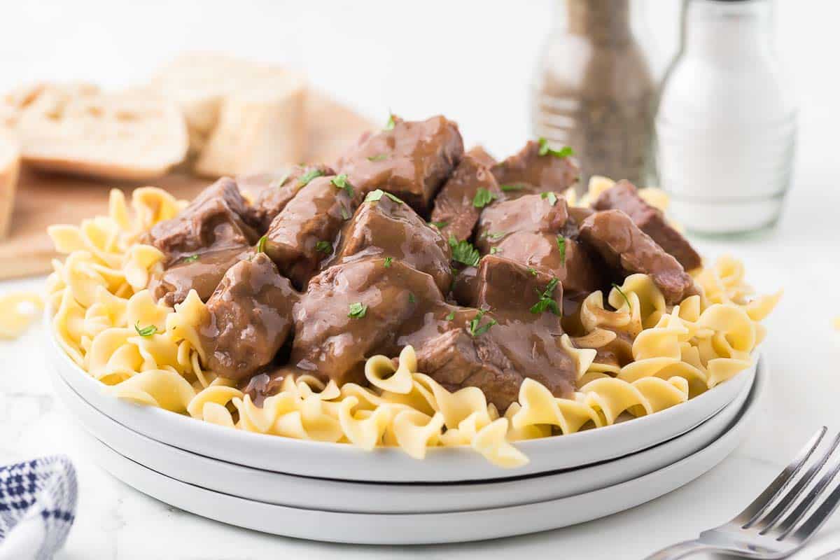 A plate of beef tips in brown gravy served over egg noodles, garnished with chopped parsley. Salt, pepper shakers, sliced bread, and a fork are in the background.