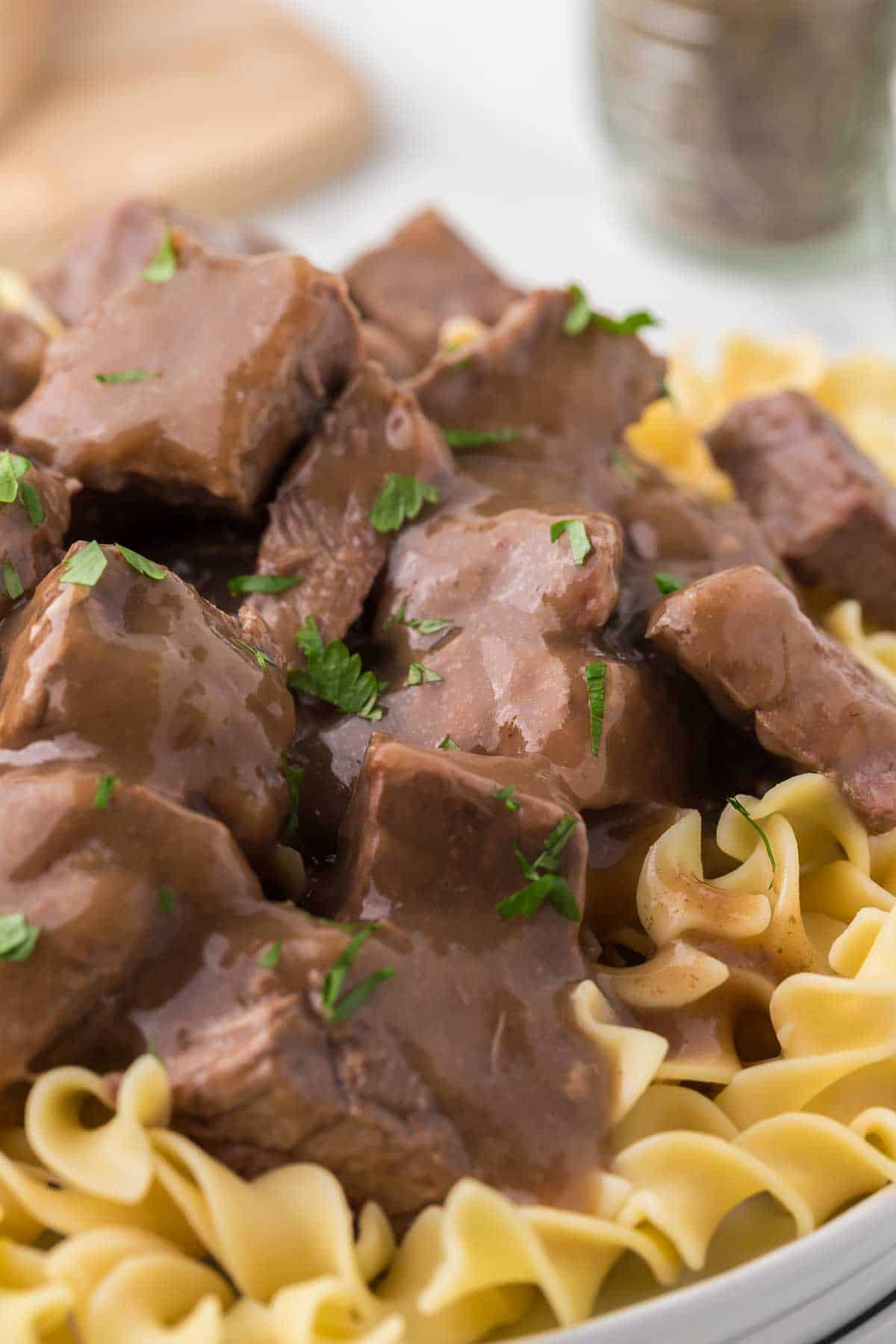 A close-up of tender beef chunks in brown gravy, served over a bed of wide egg noodles and garnished with chopped parsley.