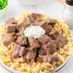 A plate of cooked egg noodles topped with chunks of beef in brown gravy, garnished with a dollop of sour cream and parsley. Peas, sliced bread, and a salt shaker are in the background.