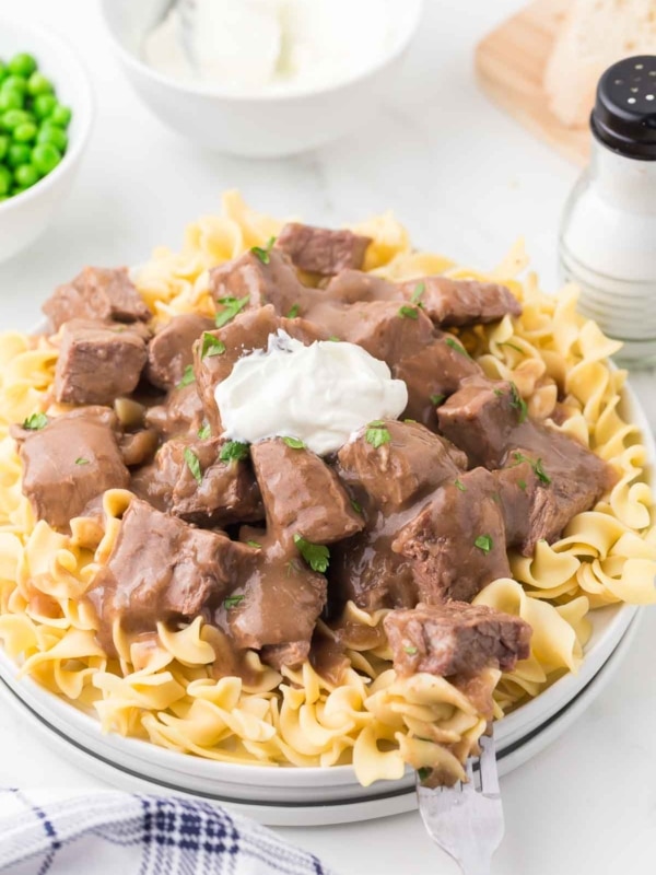 A plate of cooked egg noodles topped with chunks of beef in brown gravy, garnished with a dollop of sour cream and parsley. Peas, sliced bread, and a salt shaker are in the background.
