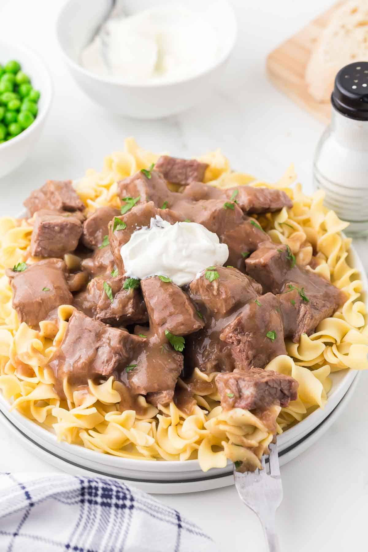A plate of cooked egg noodles topped with chunks of beef in brown gravy, garnished with a dollop of sour cream and parsley. Peas, sliced bread, and a salt shaker are in the background.