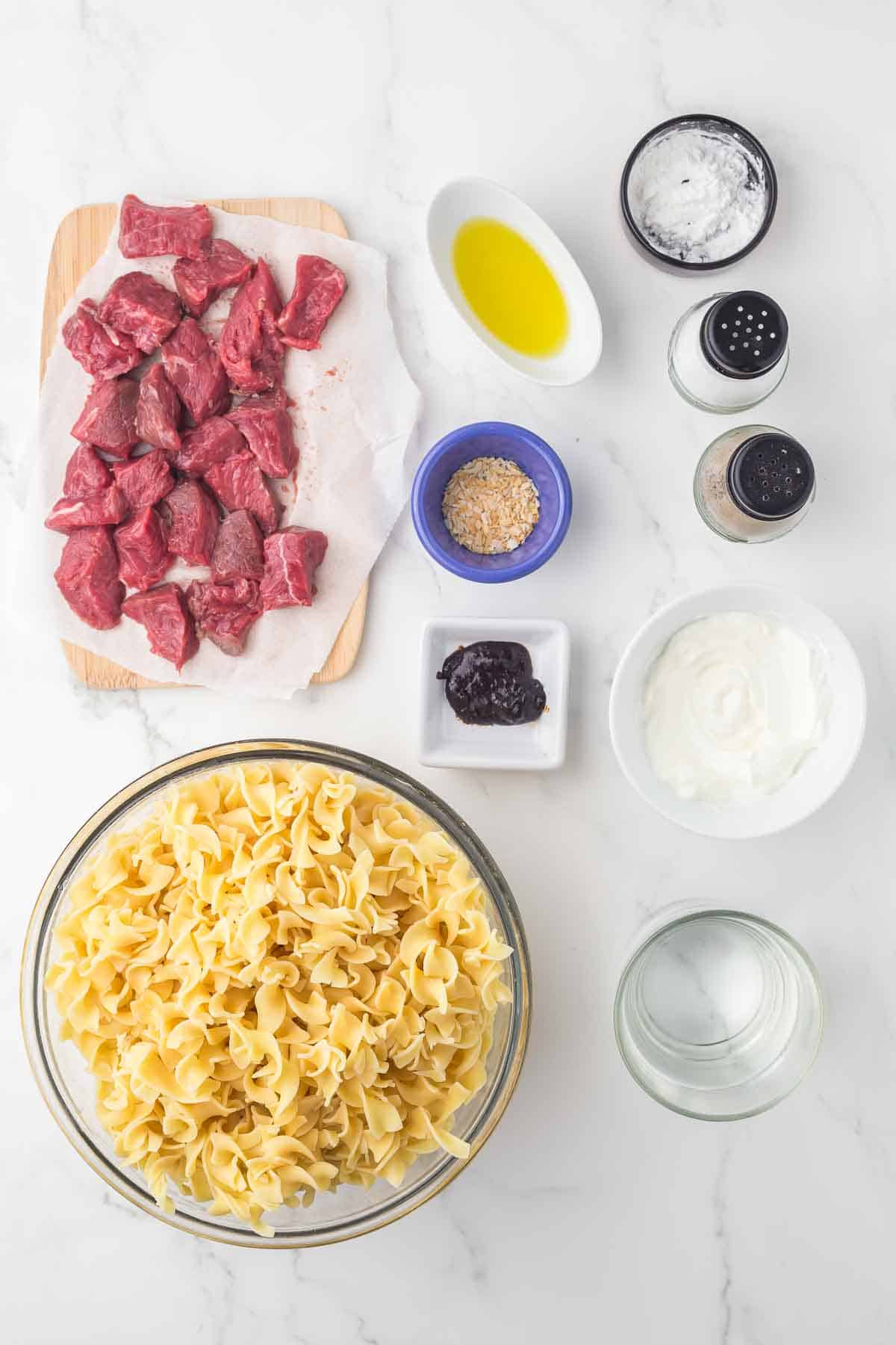 Top-down view of ingredients for beef stroganoff: raw beef chunks, uncooked egg noodles, olive oil, seasonings, sour cream, beef bouillon, flour, and water arranged in bowls on a white marble surface.