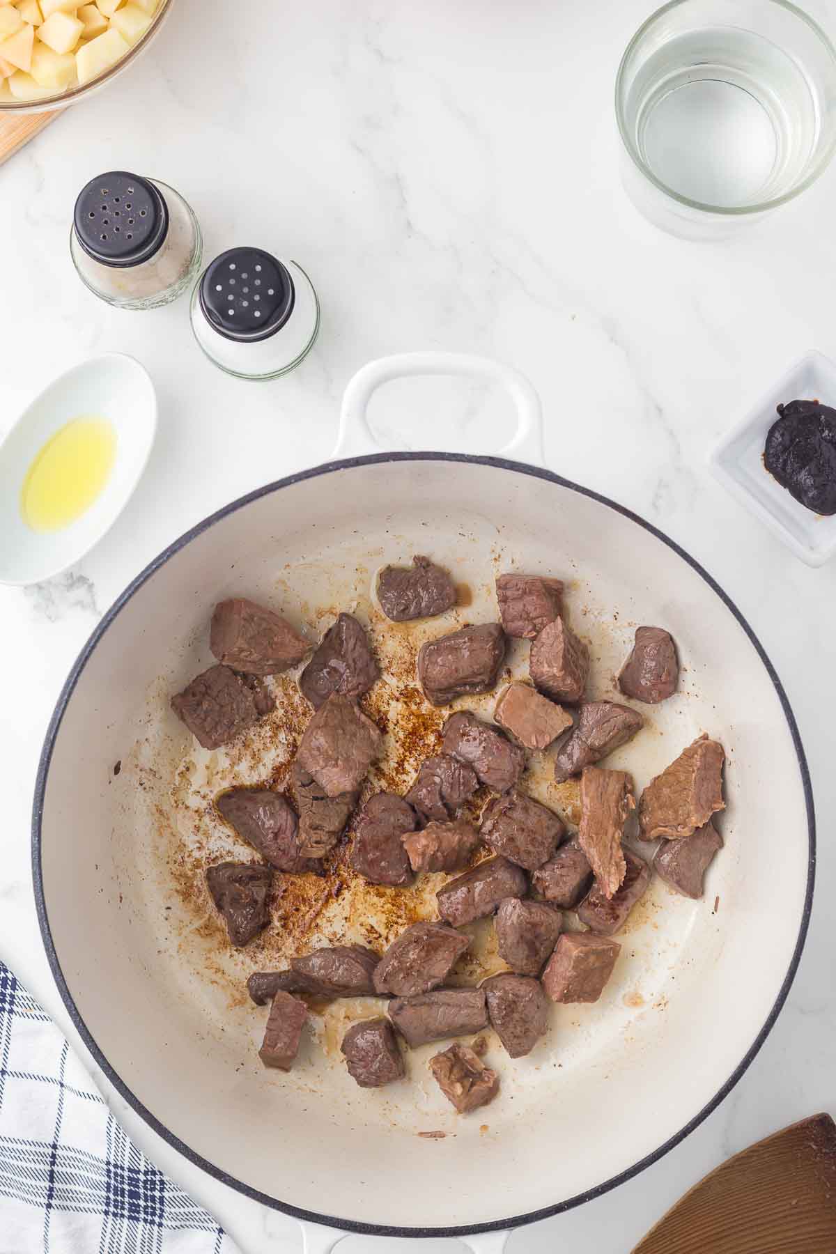 Chunks of browned beef are cooking in a white pot on a marble countertop. Surrounding the pot are bowls with oil, water, pepper and salt shakers, diced potatoes, and a dark paste on a small dish.