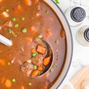 A pot of beef stew with chunks of beef, carrots, peas, and potatoes in a rich brown broth. A ladle is lifting some stew, and fresh parsley, salt, and pepper shakers are nearby on a white countertop.