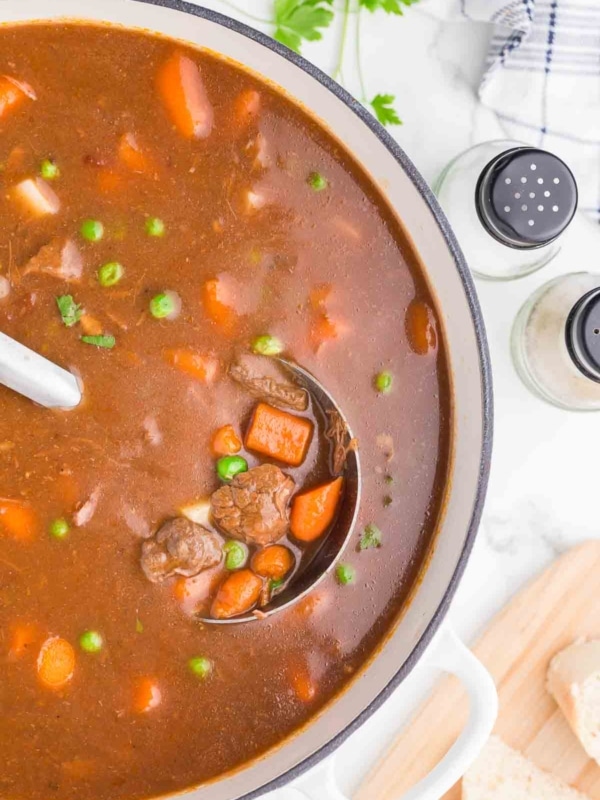A pot of beef stew with chunks of beef, carrots, peas, and potatoes in a rich brown broth. A ladle is lifting some stew, and fresh parsley, salt, and pepper shakers are nearby on a white countertop.
