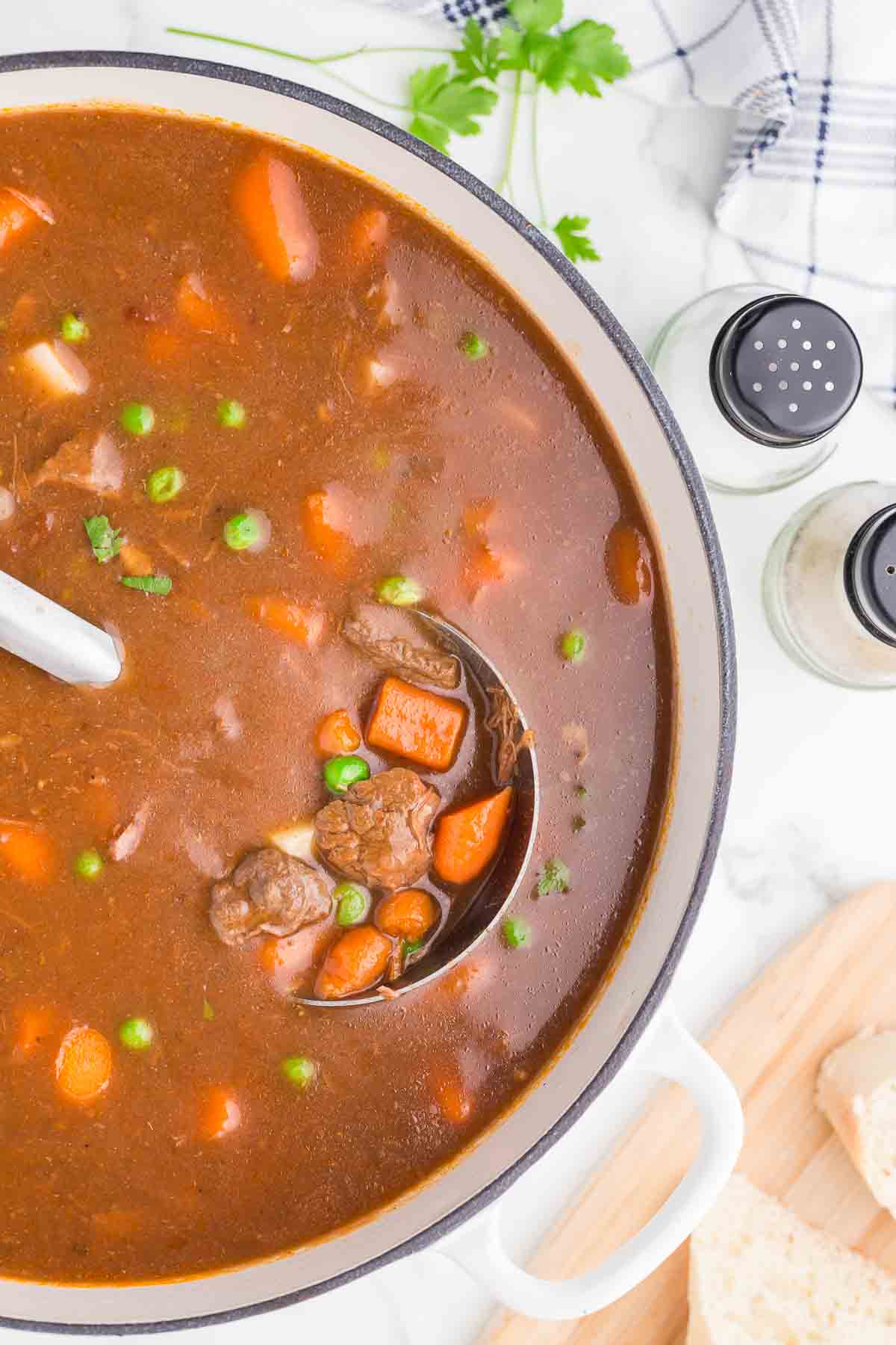 A pot of beef stew with chunks of beef, carrots, peas, and potatoes in a rich brown broth. A ladle is lifting some stew, and fresh parsley, salt, and pepper shakers are nearby on a white countertop.