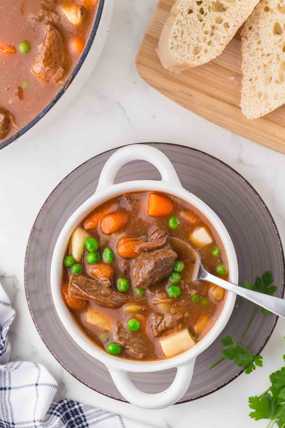 A bowl of beef stew with chunks of beef, carrots, potatoes, and green peas in a rich broth, served on a gray plate with a spoon. Slices of bread are on a wooden board nearby.