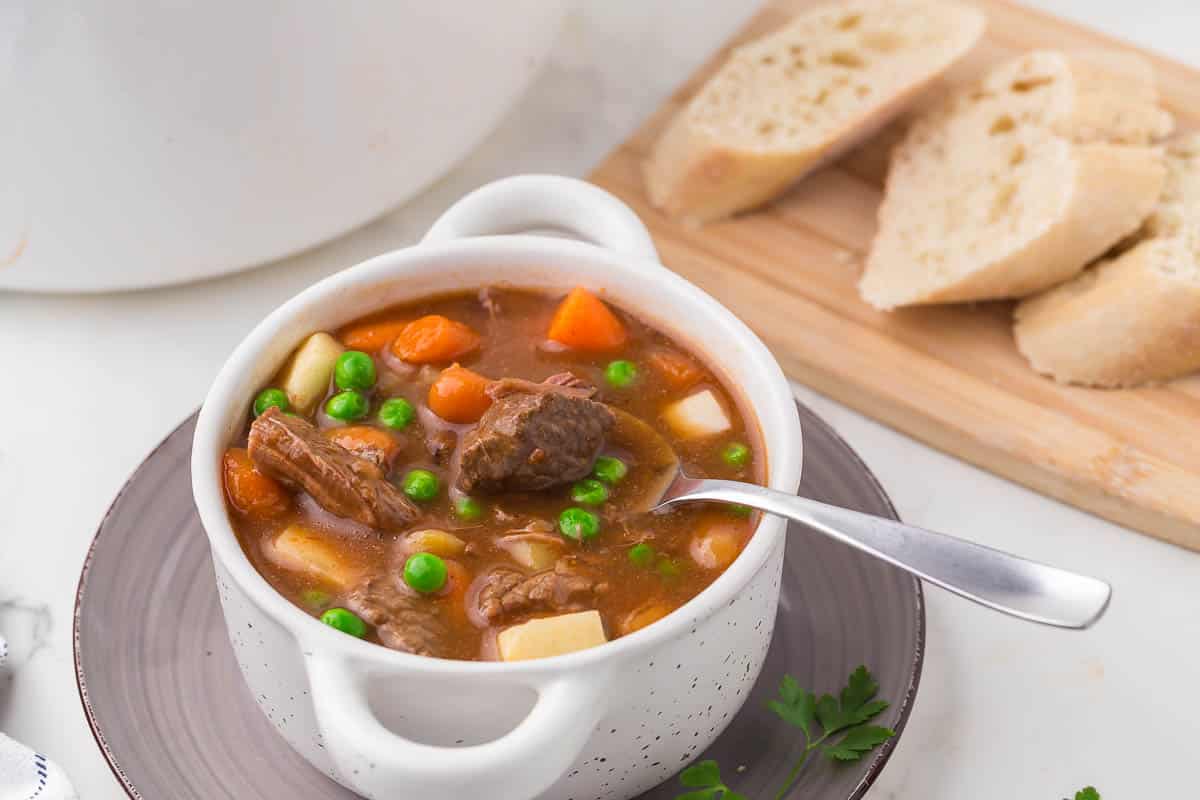 A white bowl of beef stew with peas, carrots, potatoes, and chunks of beef, sits on a gray plate with a spoon. Slices of bread are on a wooden board in the background.