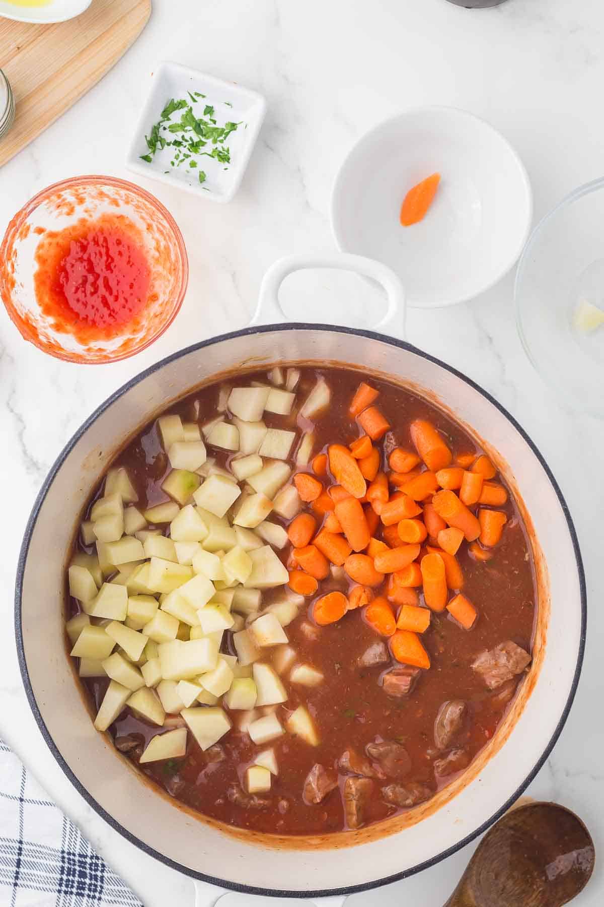 A pot of beef stew with chunks of beef, diced potatoes, and sliced carrots in a brown broth sits on a white countertop, surrounded by small bowls containing tomato sauce and chopped herbs.