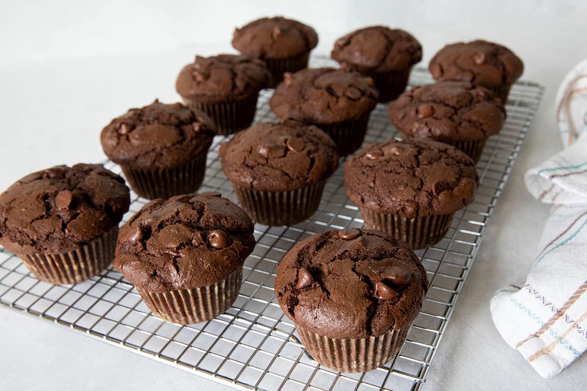 Freshly baked chocolate muffins with chocolate chips are cooling on a metal wire rack, with a light-colored towel partially visible on the right side.