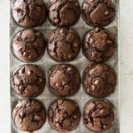 A muffin tin holds twelve freshly baked double chocolate chip muffins, each with a domed top and visible chocolate chips, set on a light-colored countertop.