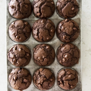 A muffin tin holds twelve freshly baked double chocolate chip muffins, each with a domed top and visible chocolate chips, set on a light-colored countertop.