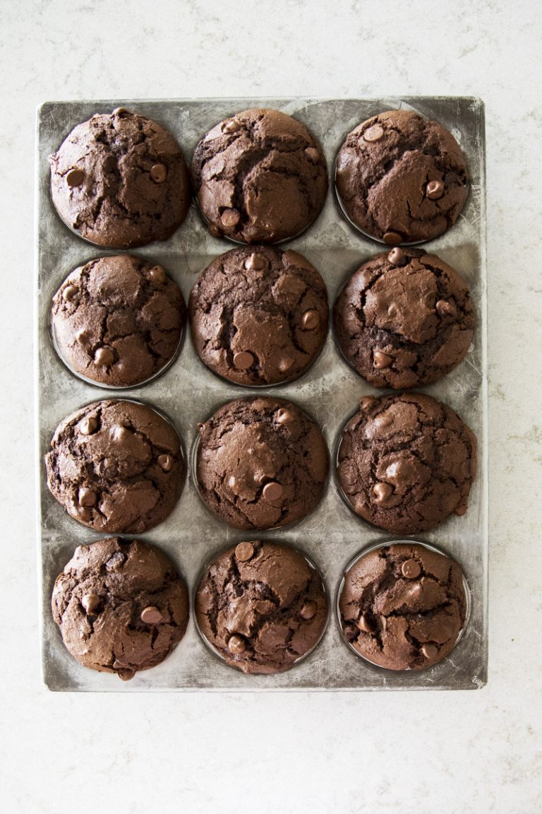 A muffin tin holds twelve freshly baked double chocolate chip muffins, each with a domed top and visible chocolate chips, set on a light-colored countertop.