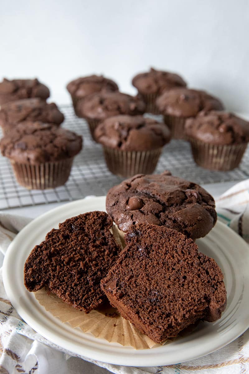 A plate with a whole chocolate muffin and a halved muffin sits in front of a cooling rack filled with more chocolate muffins. The muffins are dark and studded with chocolate chips.