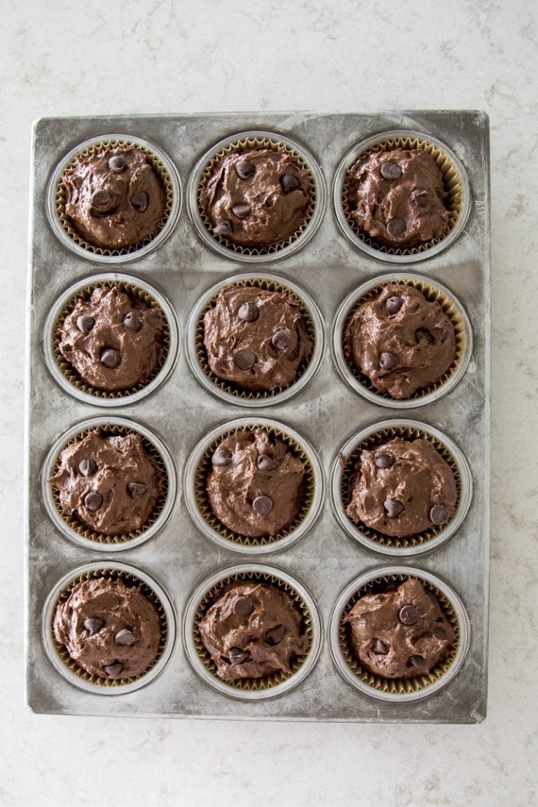 A muffin tin with twelve cups, each filled with chocolate muffin batter and topped with chocolate chips, sits on a light-colored countertop.