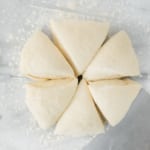 Overhead view of six triangular pieces of raw dough arranged in a circle on a floured surface, with a metal dough scraper nearby.