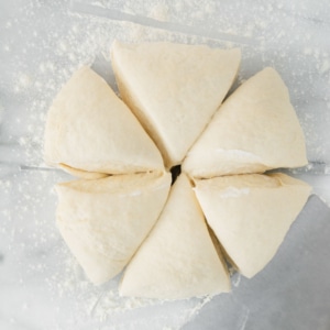 Overhead view of six triangular pieces of raw dough arranged in a circle on a floured surface, with a metal dough scraper nearby.