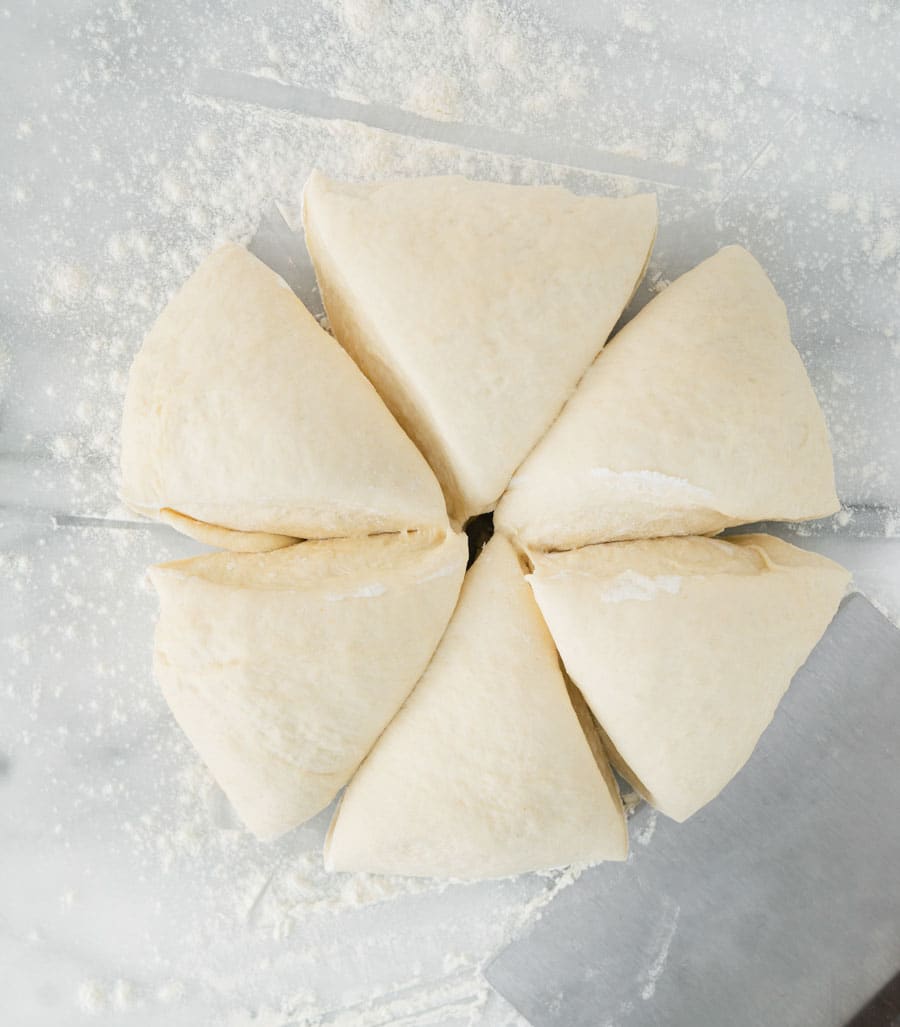 Overhead view of six triangular pieces of raw dough arranged in a circle on a floured surface, with a metal dough scraper nearby.