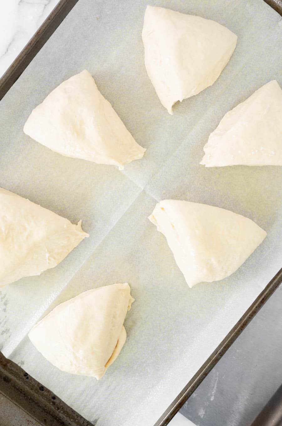 Six triangular pieces of raw dough are arranged on a parchment-lined baking sheet, ready to be baked.