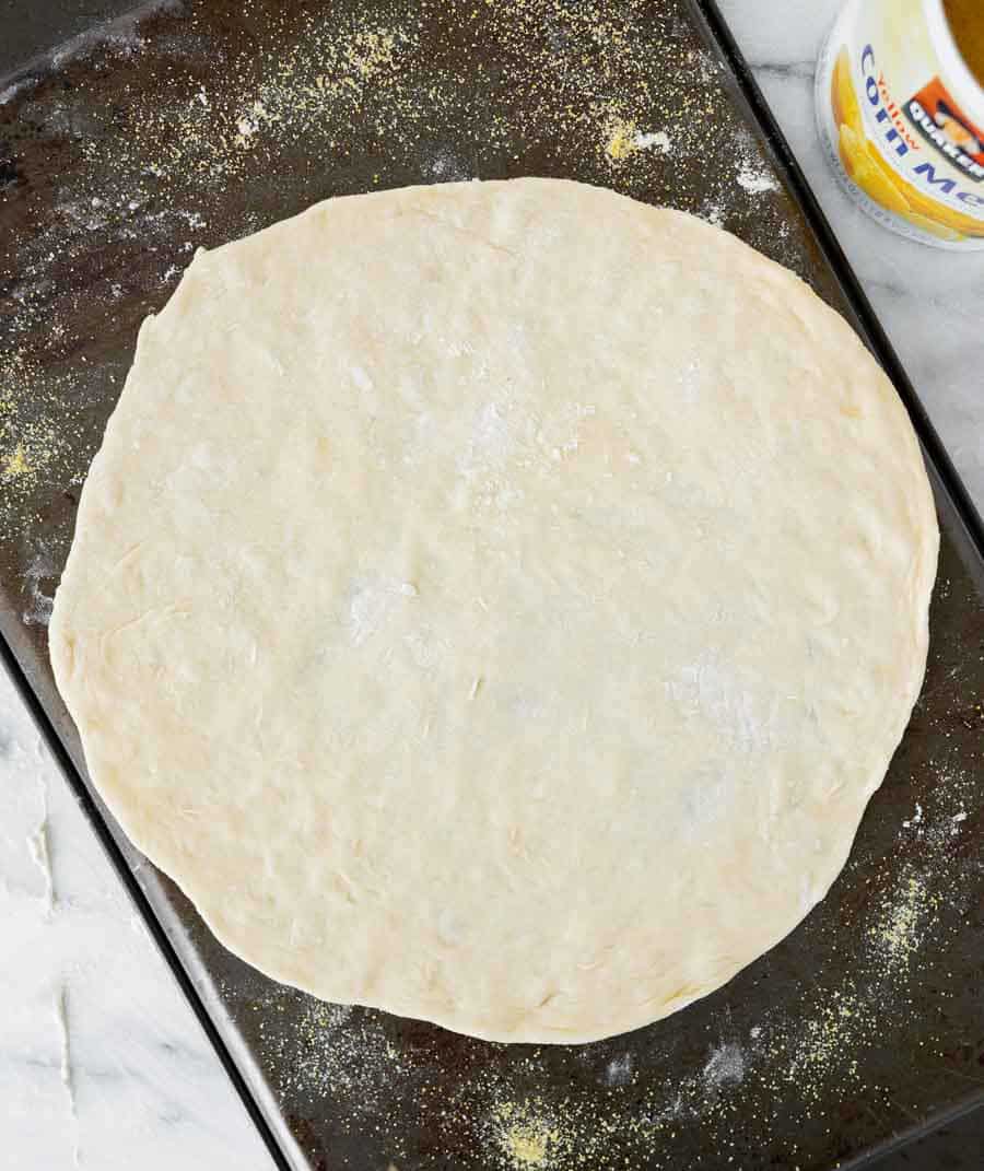 A round, uncooked pizza dough is placed on a dark baking sheet sprinkled with cornmeal. A container of cornmeal is partially visible in the top right corner.