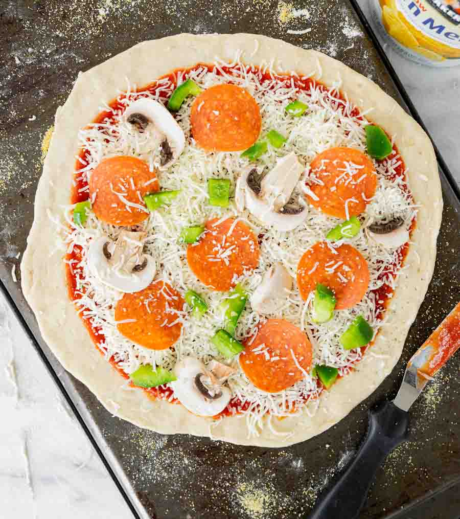 Unbaked pizza topped with shredded cheese, pepperoni slices, green bell pepper pieces, and mushroom slices on a metal baking sheet, with a pizza cutter nearby.