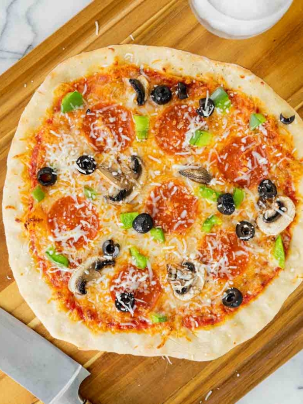 A homemade pizza topped with pepperoni, sliced mushrooms, black olives, green bell peppers, and shredded cheese sits on a wooden cutting board next to a knife and a small bowl of salt.