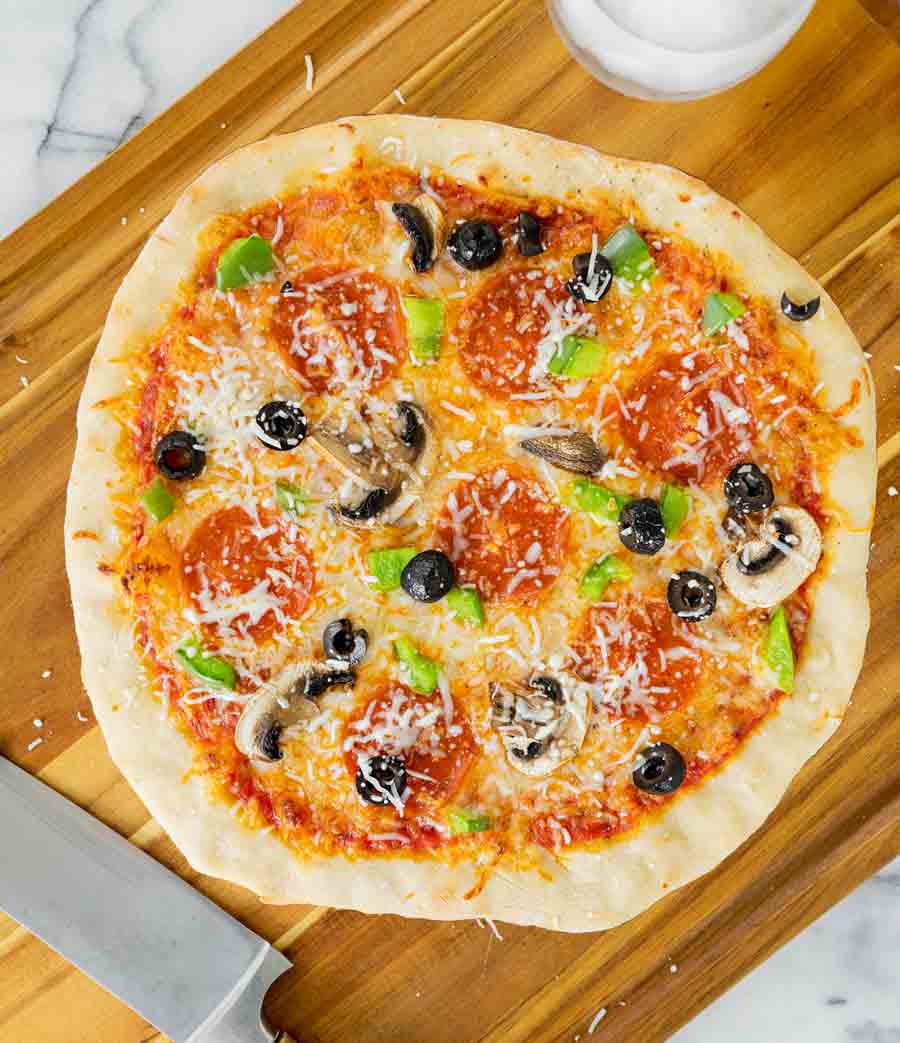 A homemade pizza topped with pepperoni, sliced mushrooms, black olives, green bell peppers, and shredded cheese sits on a wooden cutting board next to a knife and a small bowl of salt.