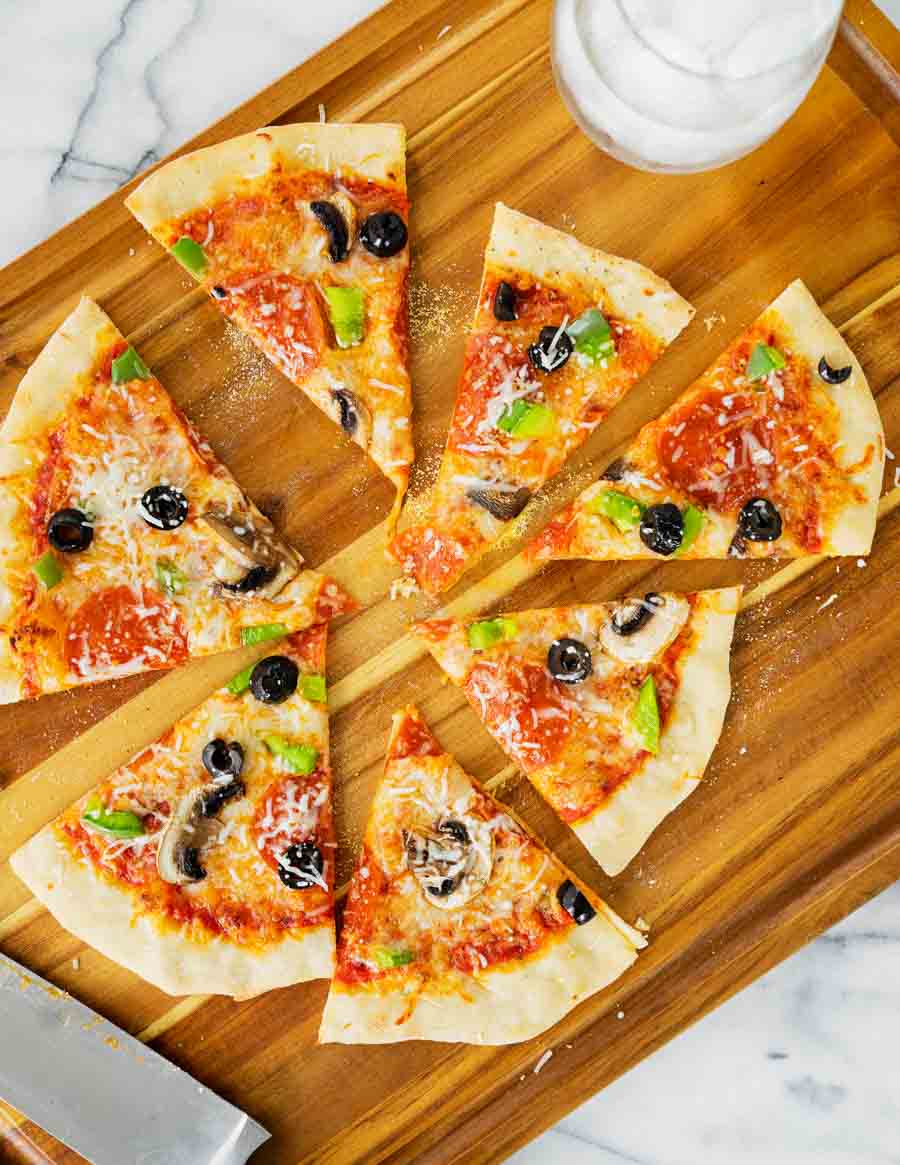 A sliced pizza with mushrooms, black olives, green peppers, pepperoni, and cheese on a wooden board, next to a knife and a glass of water.