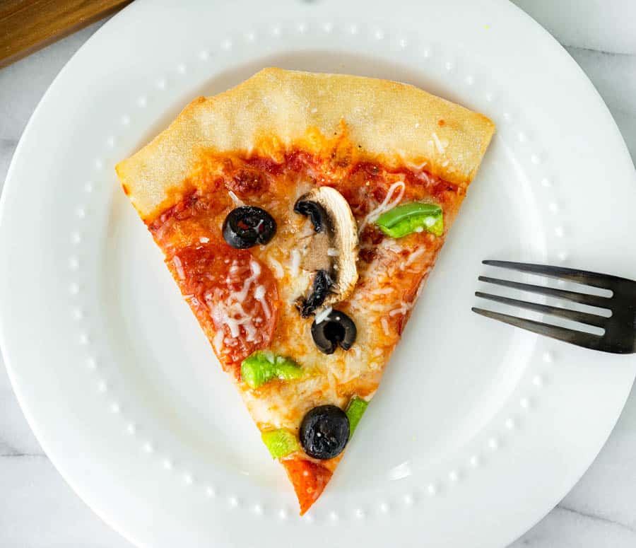 A slice of pizza on a white plate, topped with pepperoni, shredded cheese, green peppers, black olives, and a mushroom. A black fork is placed to the right of the plate.