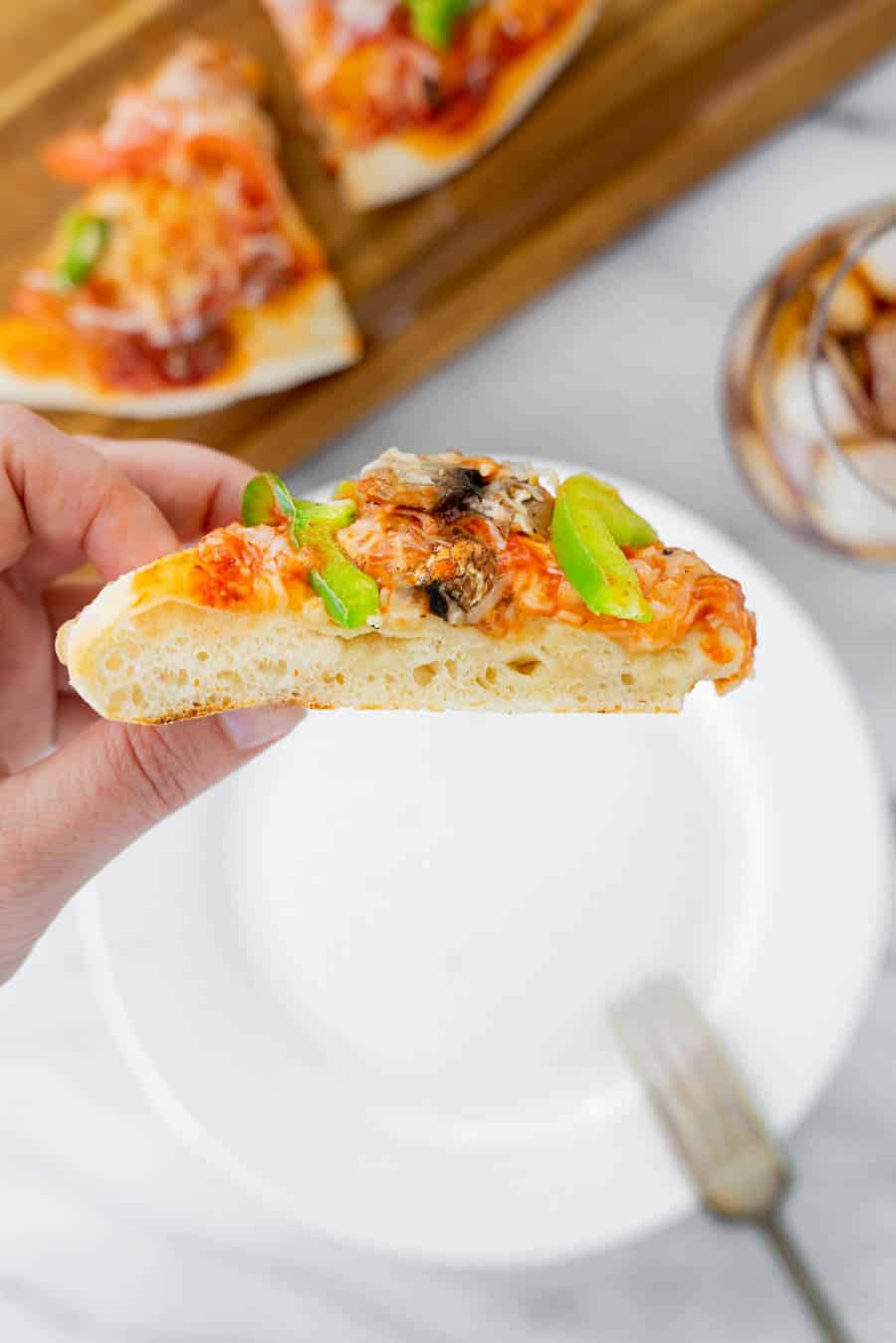 A hand holds up a slice of pizza topped with cheese, mushrooms, and green peppers. A wooden board with more pizza, a white plate, a fork, and a glass of soda are visible in the background.
