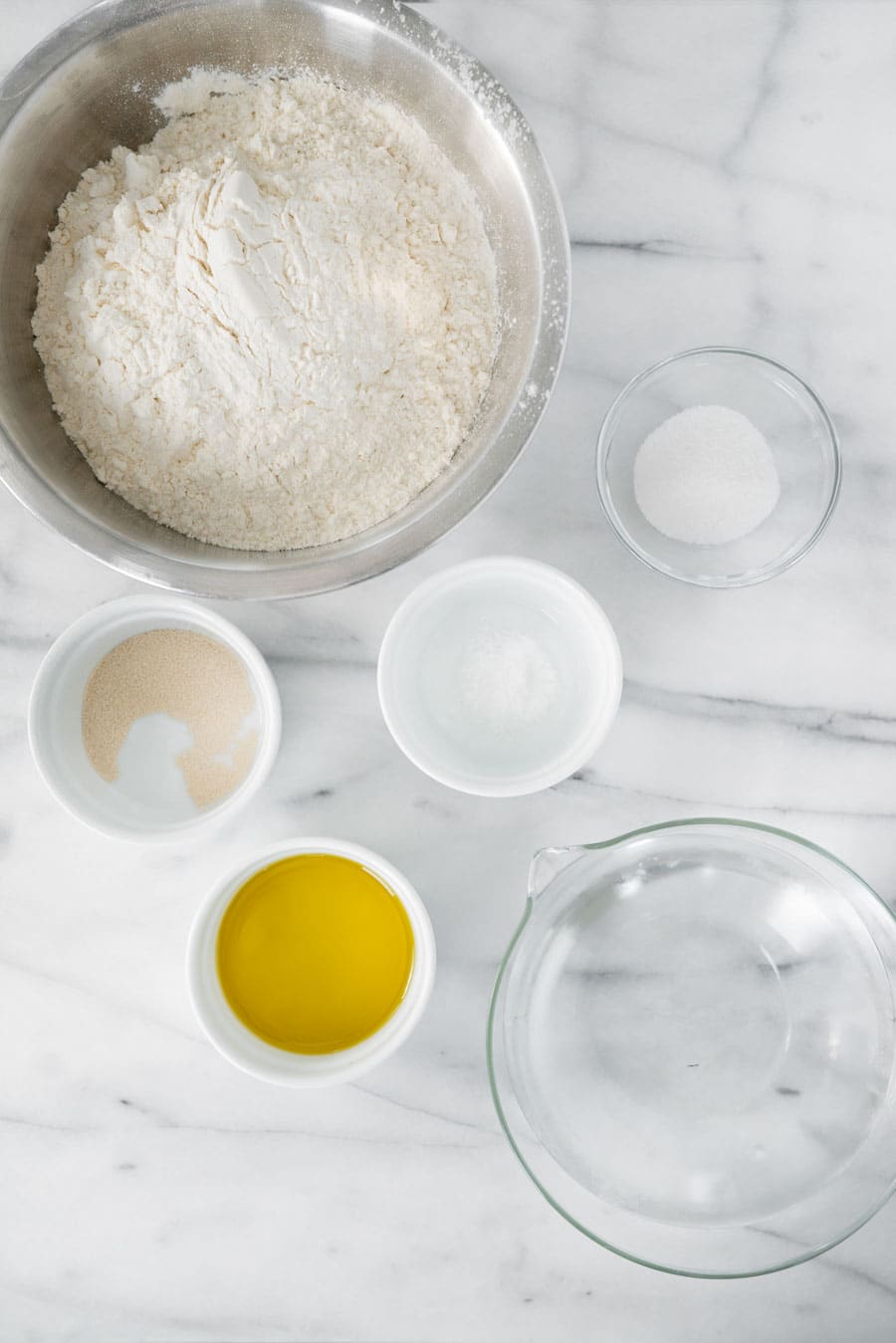 A marble countertop with bowls containing flour, sugar, instant yeast, salt, olive oil, and a glass measuring cup filled with water, arranged neatly for baking preparation.
