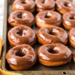 A baking tray filled with freshly glazed chocolate donuts, arranged in rows. A yellow cloth is partially visible in the bottom left corner.